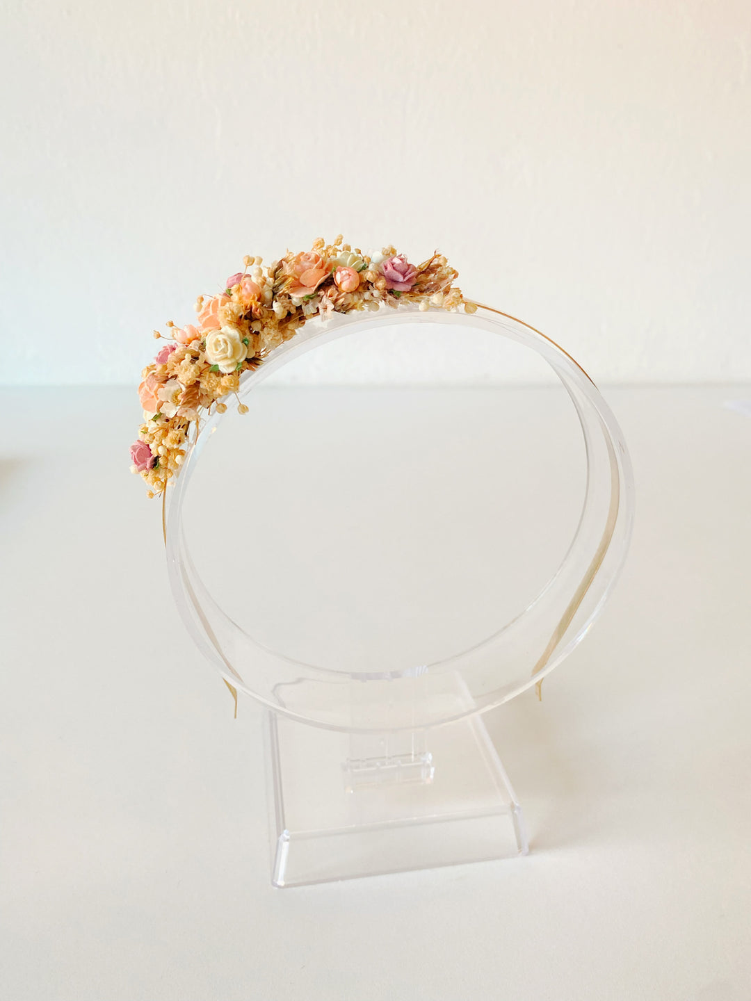 A delicate floral crown with pastel flowers and green leaves, displayed on a clear acrylic stand against a plain white background.