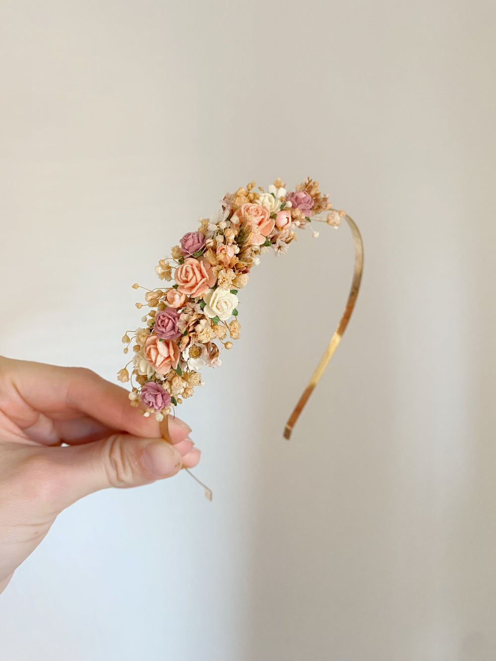 A hand holds a delicate floral headband featuring small fabric roses in peach, pink, and cream, accented with tiny white baby's breath and a gold-tone band, set against a plain light beige background.