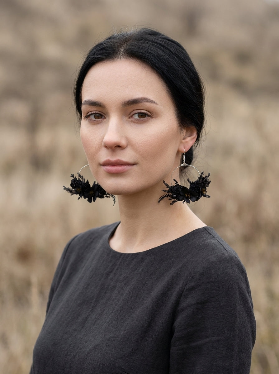 A woman with dark hair pulled back wears large black feathered hoop earrings with silver-toned metal rings, photographed outdoors against a blurred dry grass background.