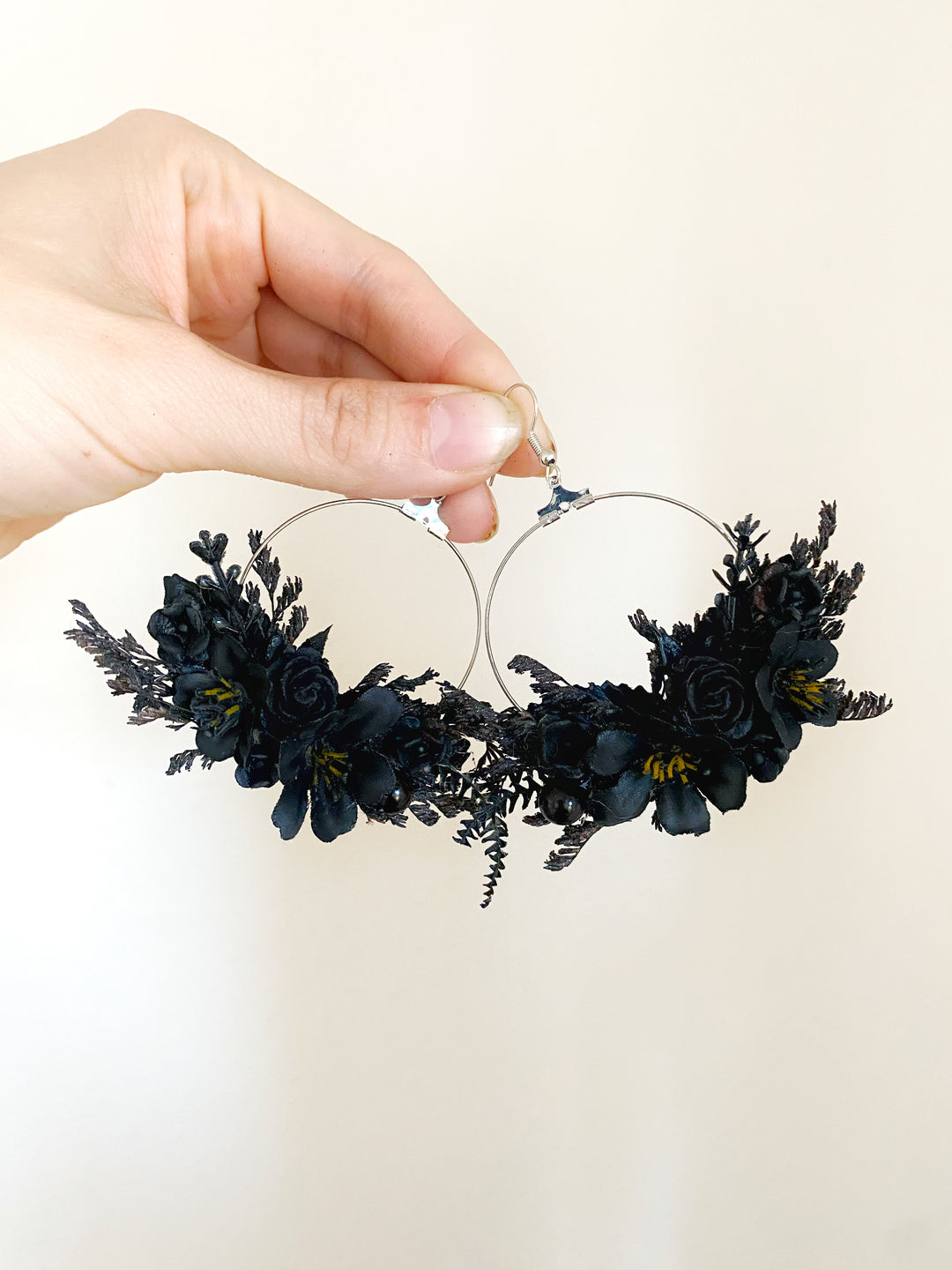 A hand holding a pair of silver hoop earrings adorned with black dried flowers and dark green foliage against a plain off-white background.