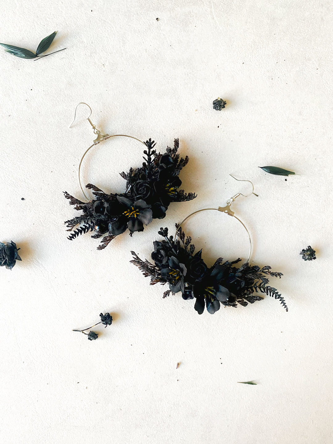 A pair of large silver-tone hoop earrings adorned with black flowers and foliage, arranged on a light beige textured surface with scattered dried botanicals.