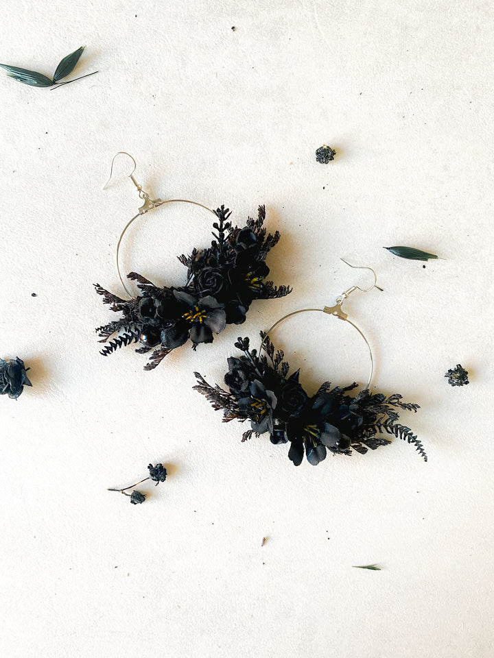 A pair of large silver-tone hoop earrings adorned with black flowers and foliage, arranged on a light beige textured surface with scattered dried botanicals.
