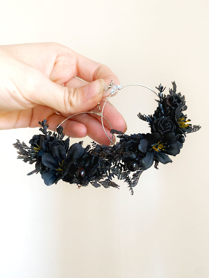 A hand holding a black floral hoop earring with silver-tone metal hoops and dark artificial flowers, set against a plain light beige background.