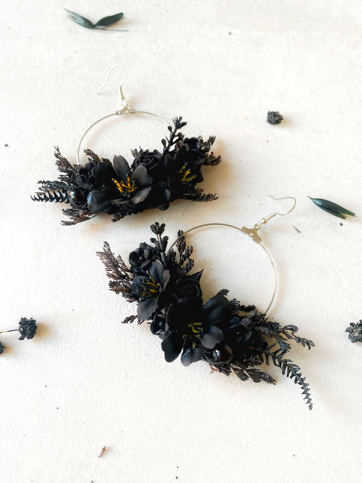 A pair of large silver hoop earrings adorned with black flowers and dark green foliage, displayed on a textured off-white surface with scattered dried botanicals.