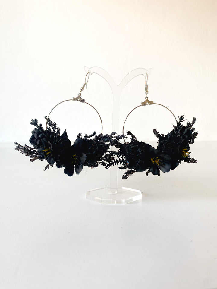 Black floral hoop earrings with silver-tone wire hoops and gold-toned hooks, displayed on a clear acrylic stand against a white background.