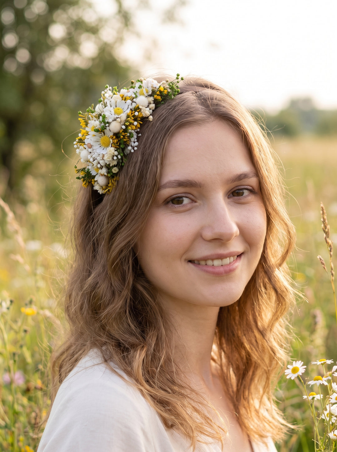 A smiling young woman outdoors in a sunlit field, wearing a floral crown made of white daisies and small yellow flowers.