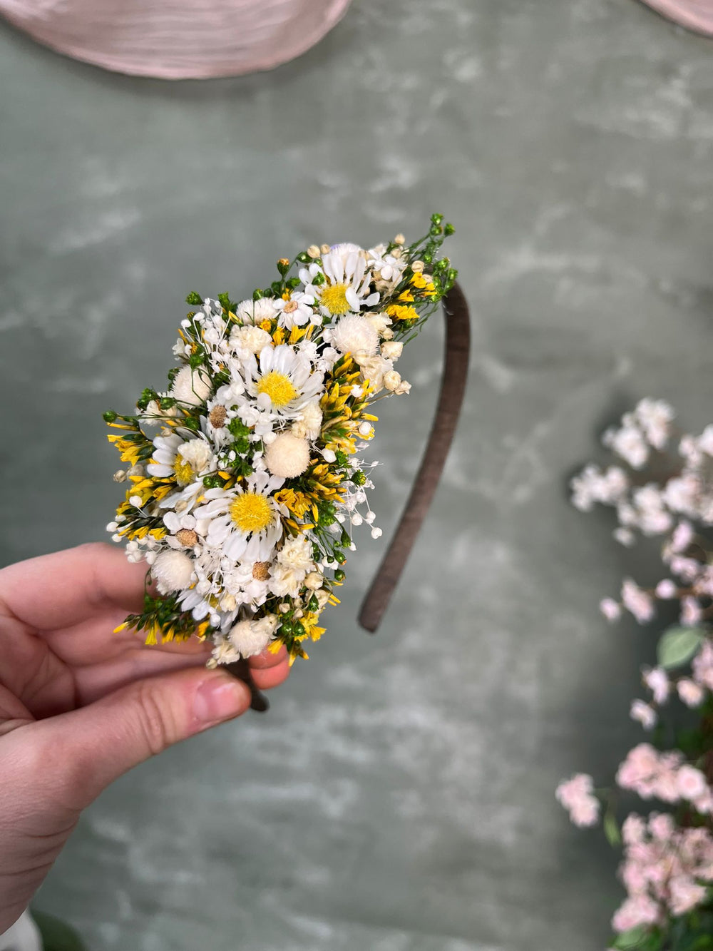 A hand holding a small floral headband decorated with white daisies, yellow wildflowers, and green leaves, set against a textured gray background.