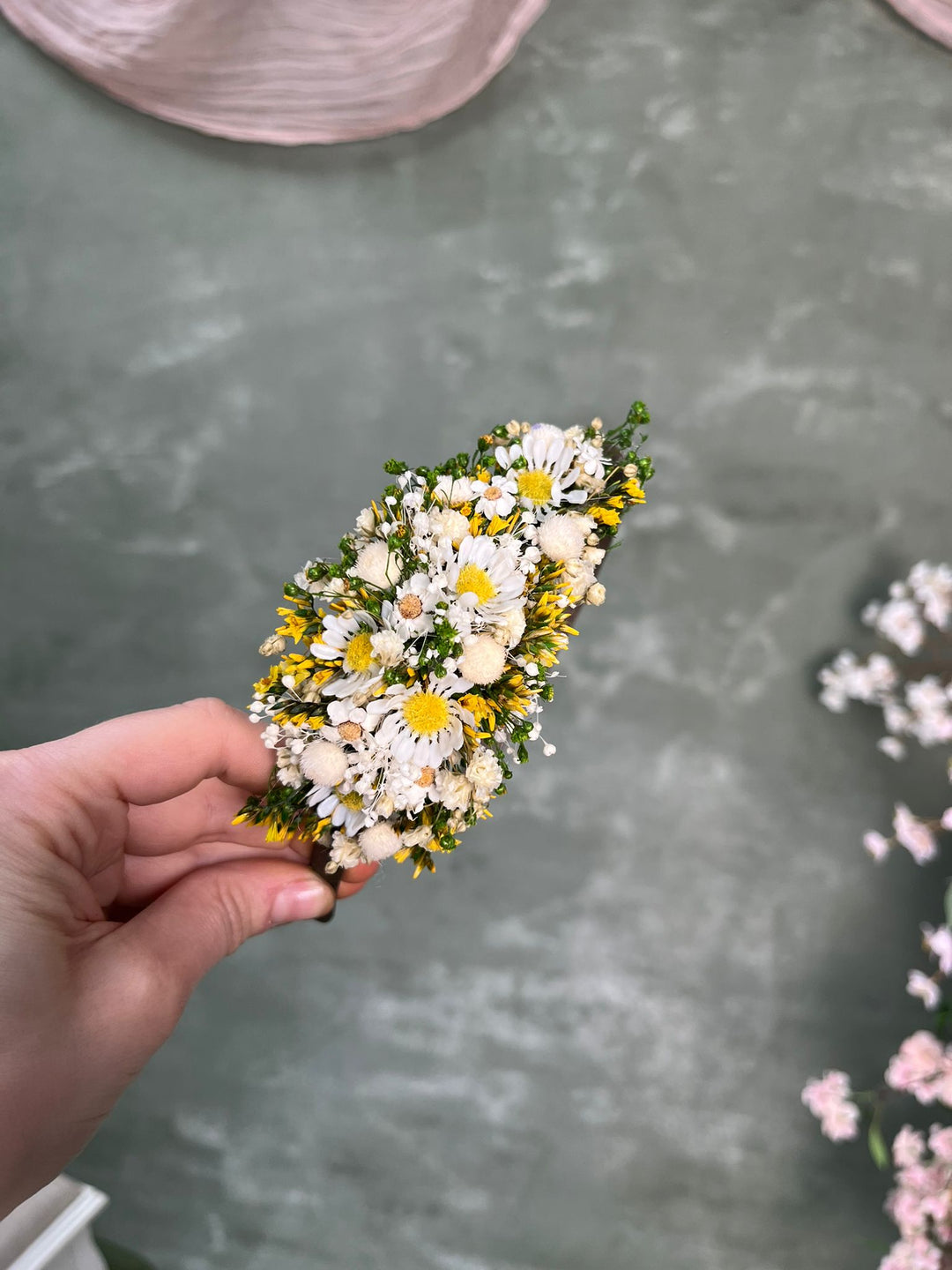A hand holding a small flower arrangement featuring white and yellow daisies with green foliage, set against a textured gray background with blurred pink flowers.