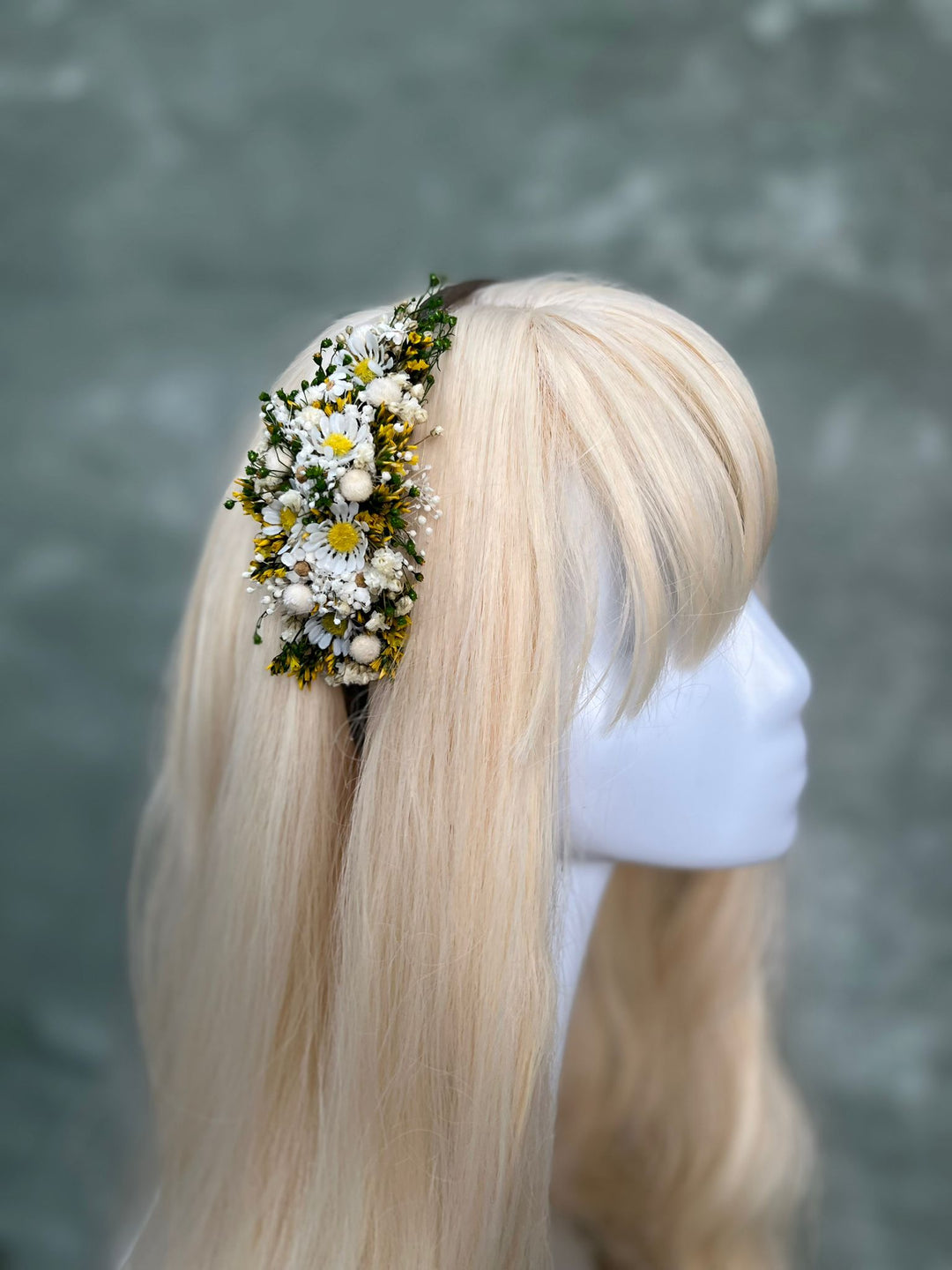 A blonde wig displayed on a white mannequin head, featuring a side-swept bang and a floral hair accessory with white daisies and green leaves on the side, set against a textured gray background.