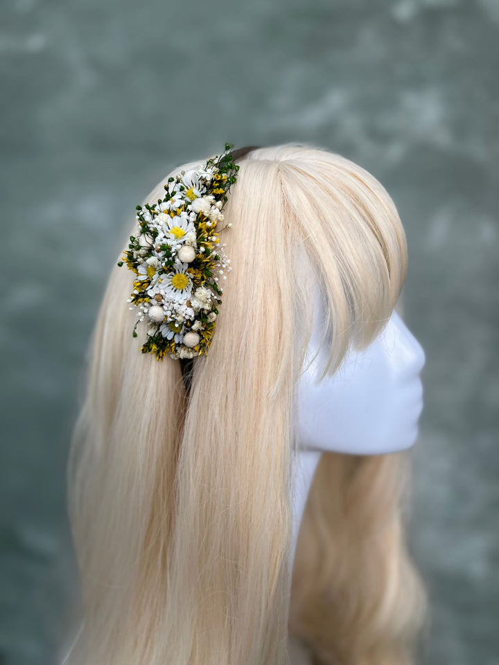 A blonde wig displayed on a white mannequin head, featuring a side-swept bang and a floral hair accessory with white daisies and green leaves on the side, set against a textured gray background.
