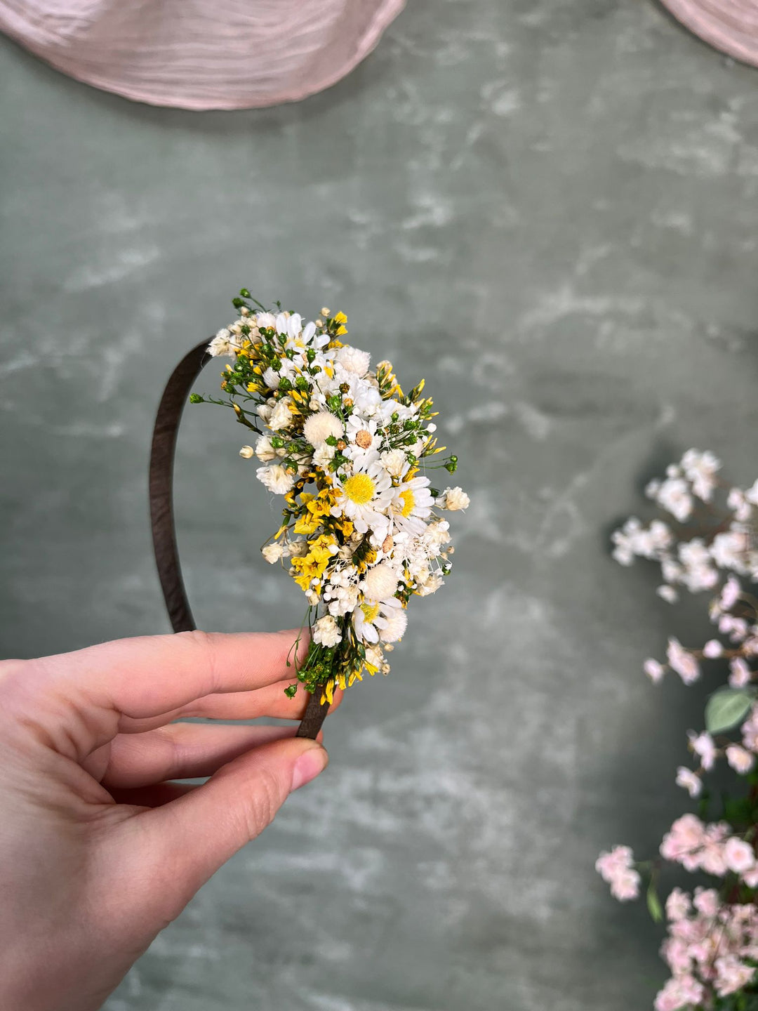 A hand holds a dark brown headband decorated with a small bouquet of white and yellow daisies and green foliage, set against a textured gray background with soft pink floral accents.