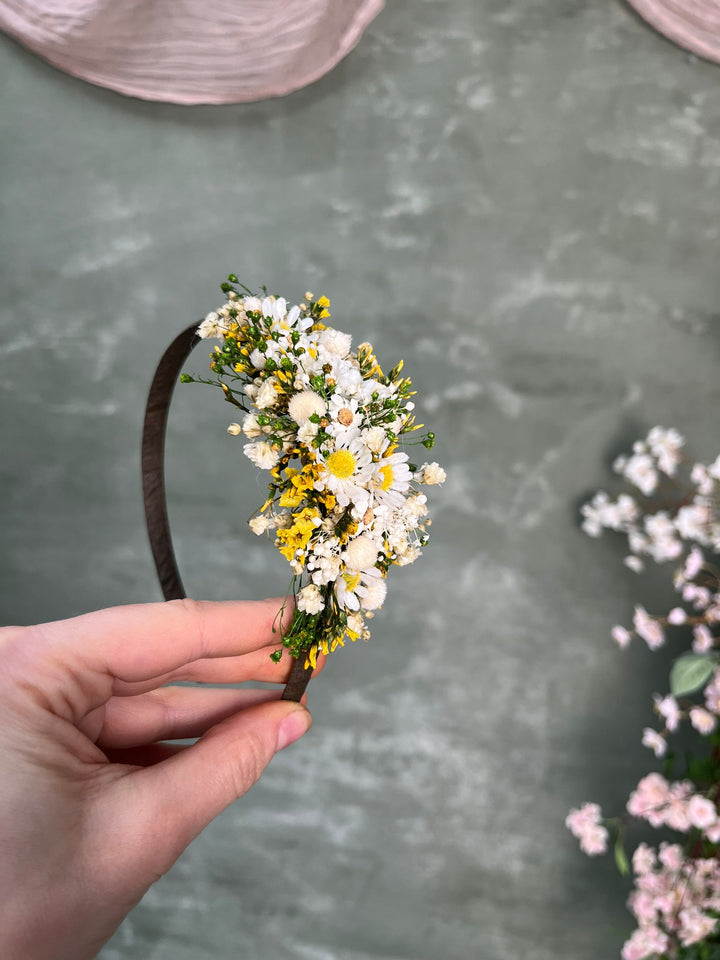 A hand holds a dark brown headband decorated with a small bouquet of white and yellow daisies and green foliage, set against a textured gray background with soft pink floral accents.