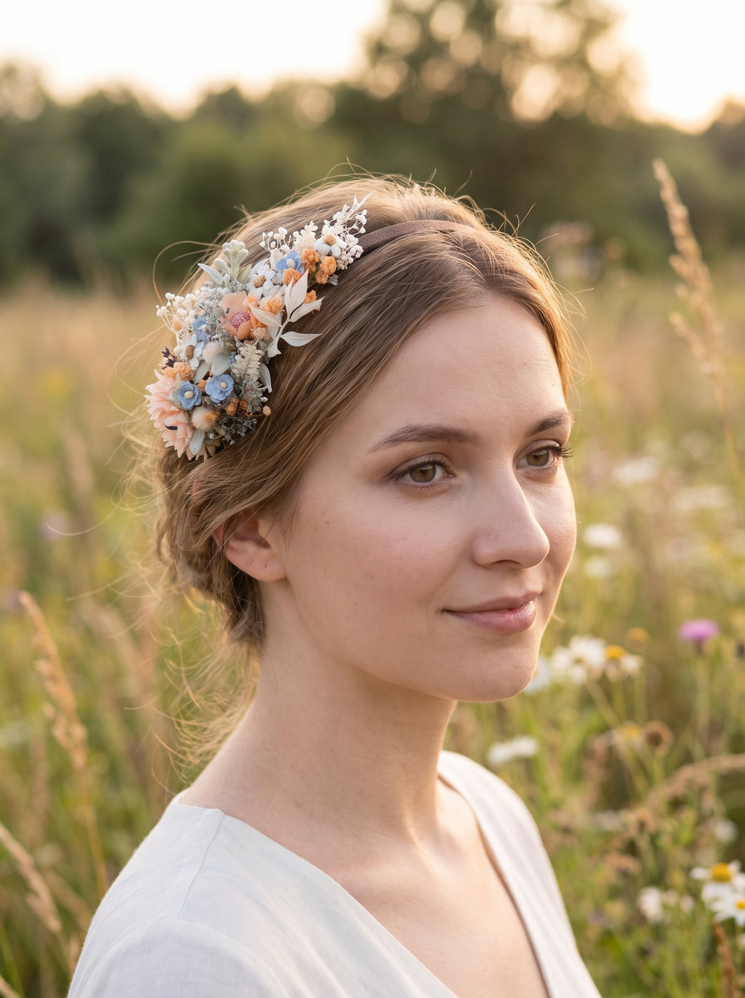 A woman in a white dress wearing a delicate floral crown with flowers in soft pastel colors, standing in a sunlit grassy field.