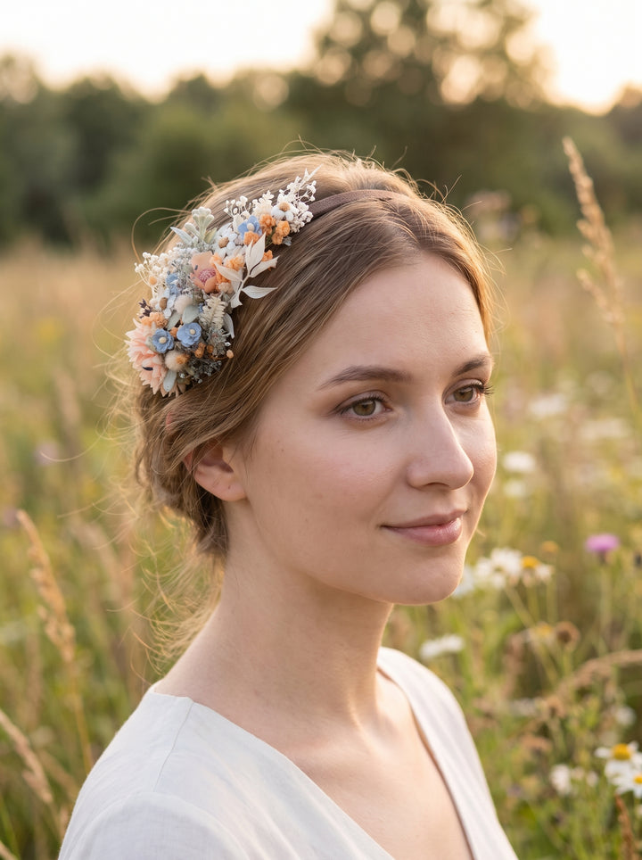 A woman in a white dress wearing a delicate floral crown with flowers in soft pastel colors, standing in a sunlit grassy field.