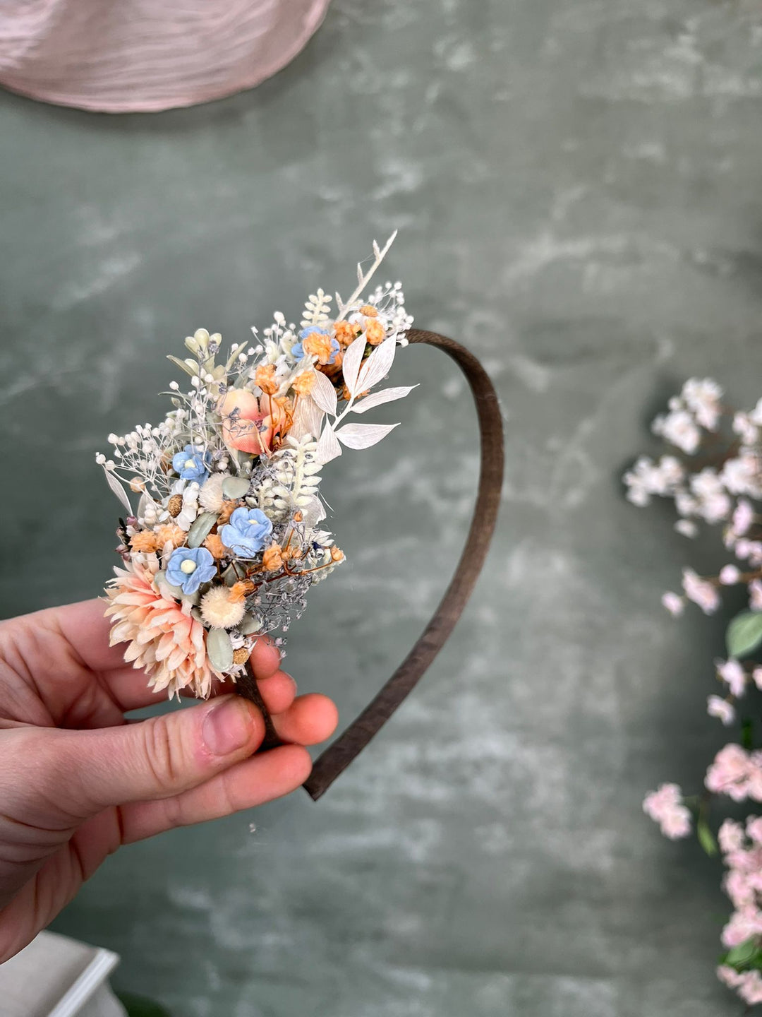 A hand holds a dark brown headband decorated with a floral arrangement in pastel peach, blue, cream, and white, set against a textured gray background.