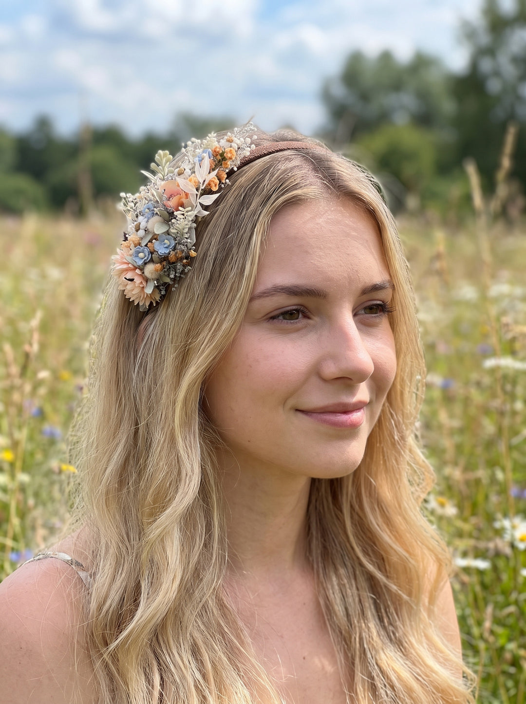 A woman with long blonde hair wearing a bohemian-style flower crown in a sunlit grassy field.