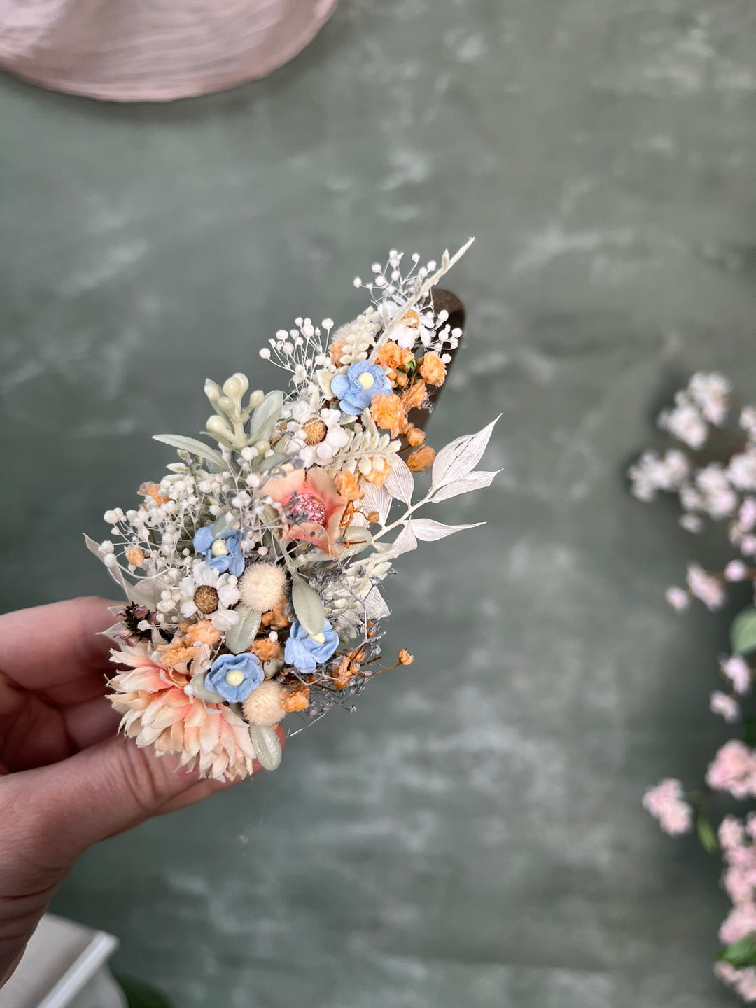 A hand holding a floral hair clip against a textured gray-green background.
