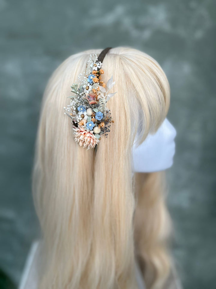 A blonde mannequin or wig display wearing a decorative hair accessory with dried flowers, small shells, and beads on a dark band, photographed against a soft gray-green textured background.