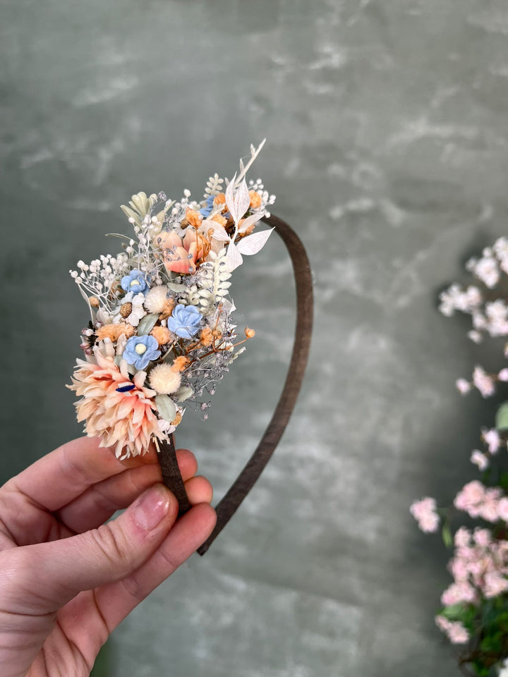 A hand holding a dark brown headband decorated with a floral arrangement in soft peach, cream, light blue, and white tones, set against a textured gray background.