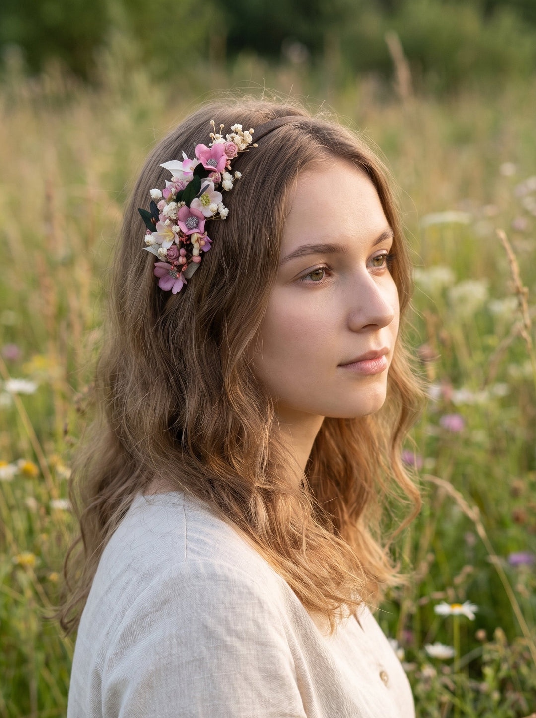 A woman with wavy light brown hair wearing a pink and white wildflower crown, standing in a sunlit grassy field.