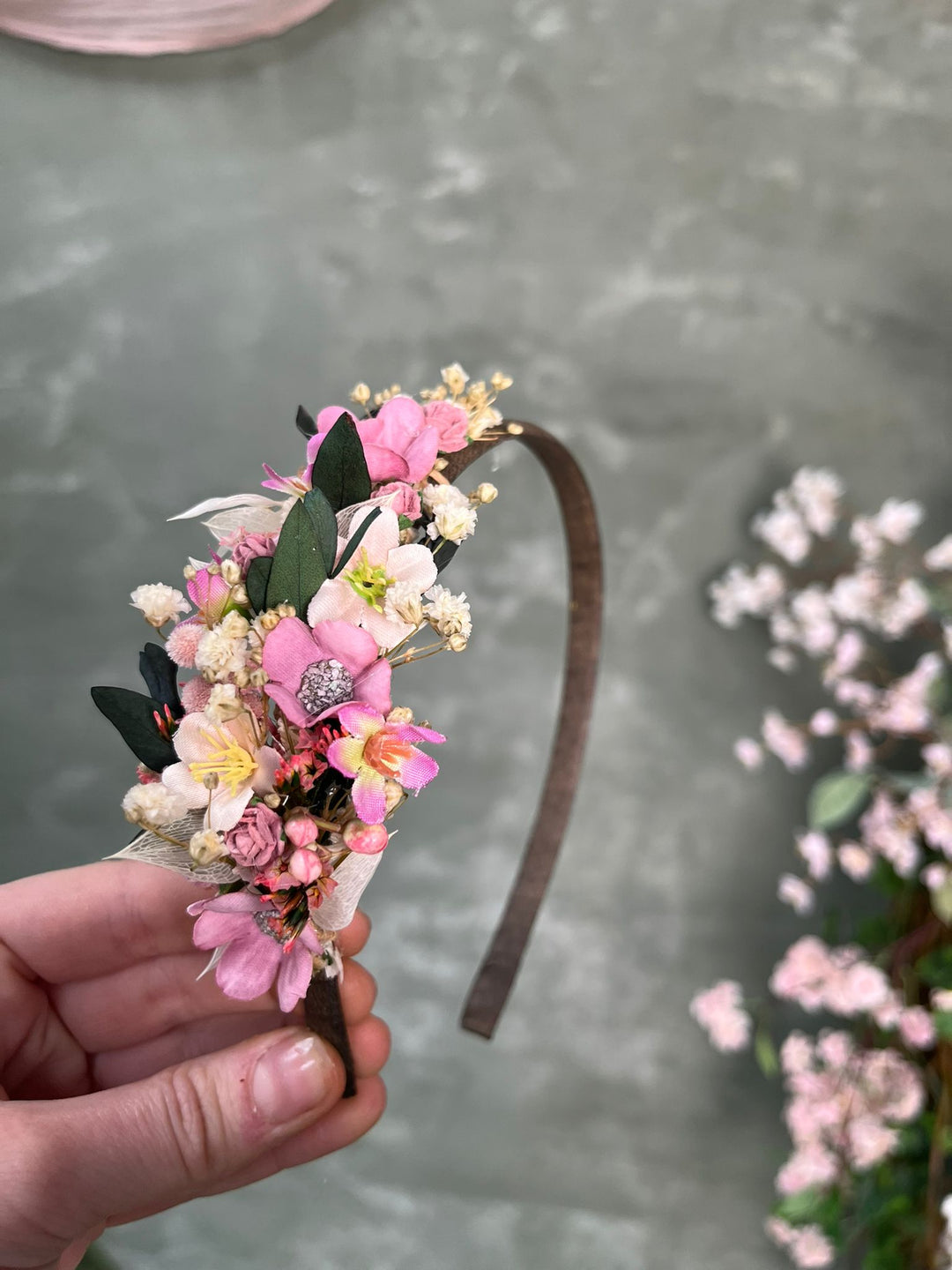 A hand holds a brown headband decorated with pink and white flowers, green leaves, against a gray textured background with blurred pink flowers.