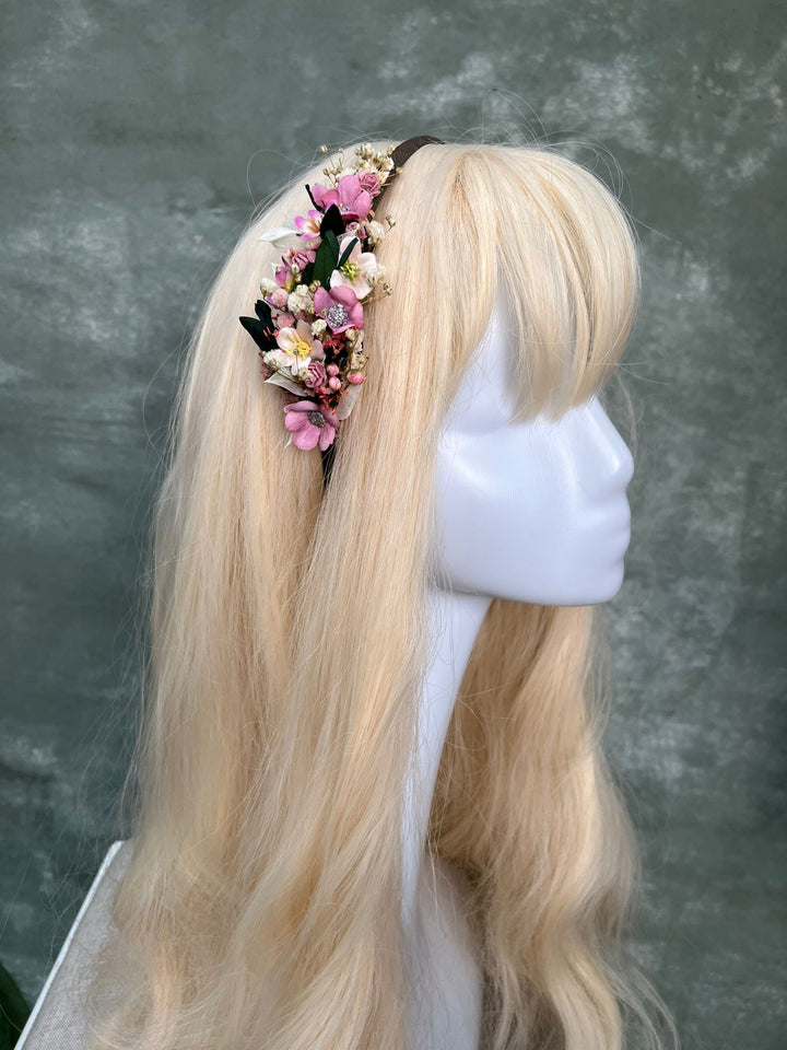 A long blonde wig with straight bangs displayed on a white mannequin head, featuring a floral headband with pink, white, and green flowers against a textured gray background.