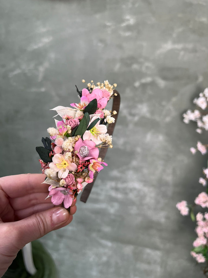 A hand holding a small floral headband decorated with pink, white, and yellow dried flowers and green leaves, set against a textured gray background.