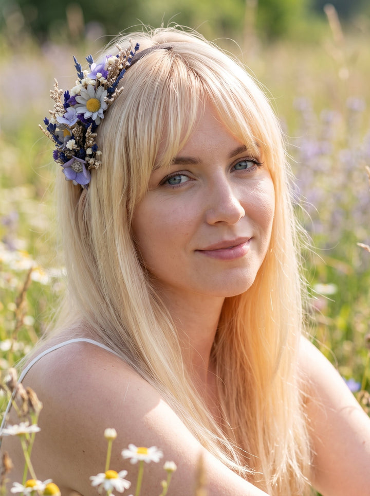 A blonde woman in a sunlit field wearing a floral hair crown made of white daisies, lavender, and small wildflowers.