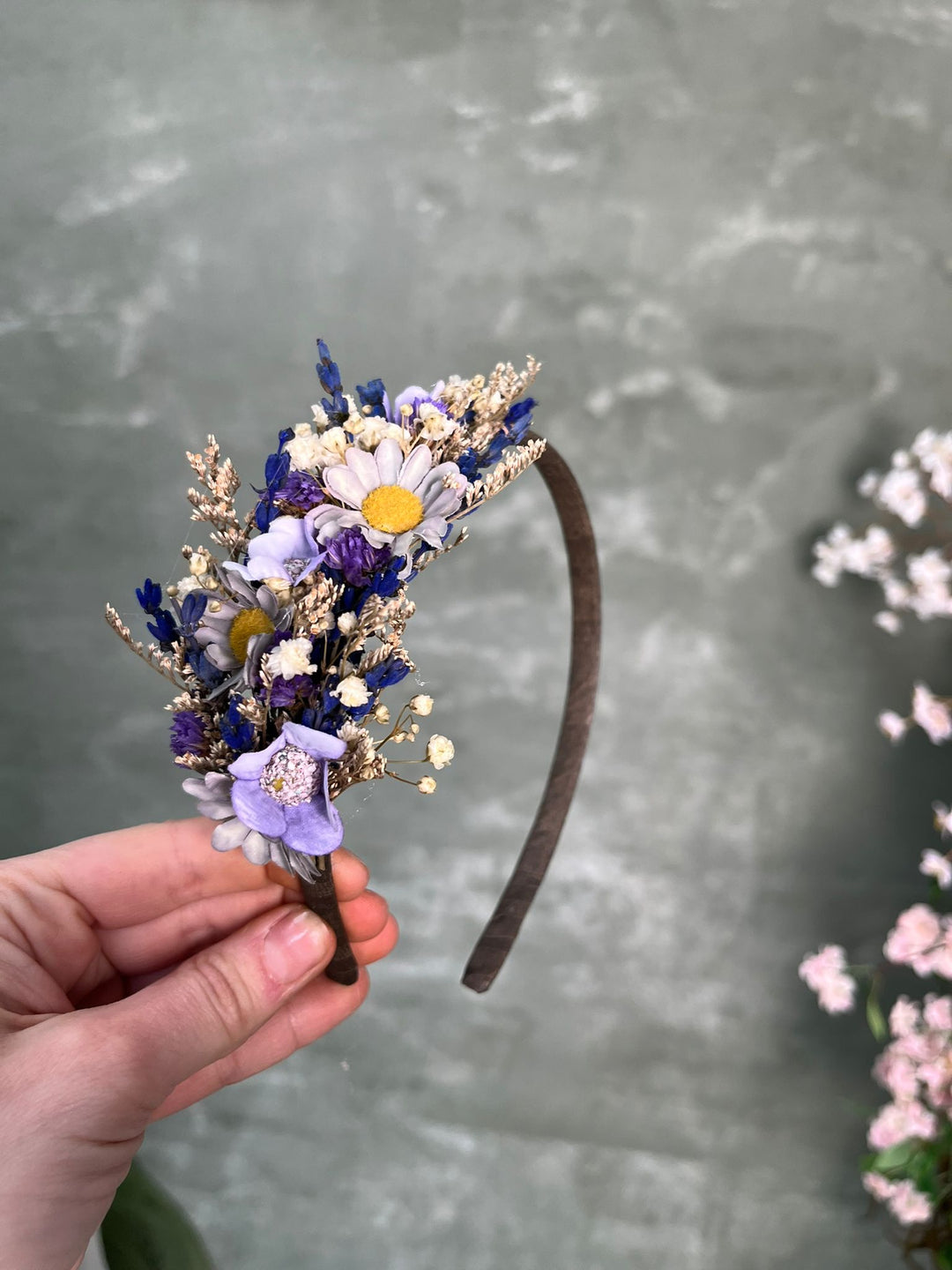 A hand holding a small flower headband with purple, white, and yellow blossoms on a dark brown band, set against a textured gray background with blurred pink flowers on the right.