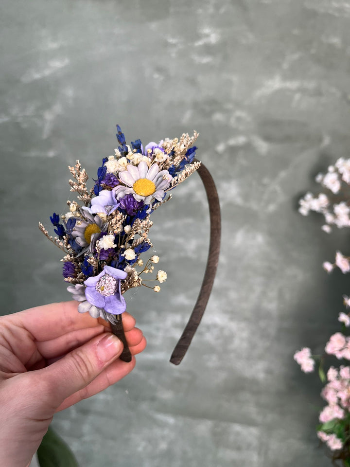 A hand holding a small flower headband with purple, white, and yellow blossoms on a dark brown band, set against a textured gray background with blurred pink flowers on the right.