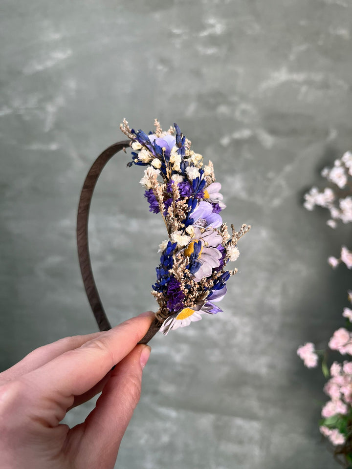 A hand holding a dark brown headband decorated with dried purple, blue, white, and yellow flowers against a textured gray background.