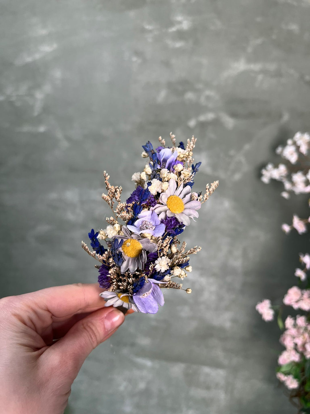 A hand holding a headband with purple, white, and yellow blossoms against a textured gray background.