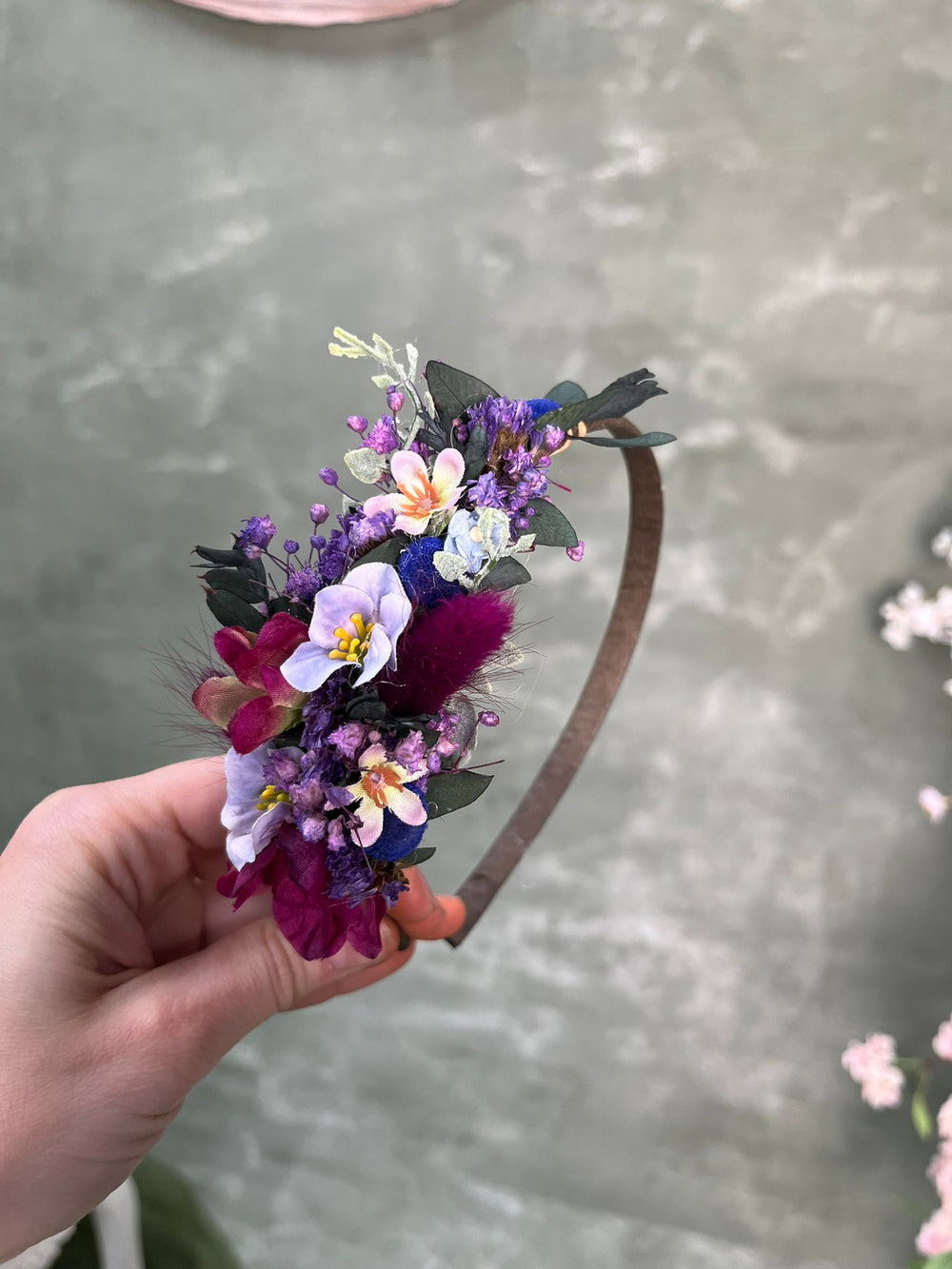A hand holding a brown headband decorated with a dense cluster of flowers in purple, magenta, white, and yellow with green foliage, set against a textured gray background.