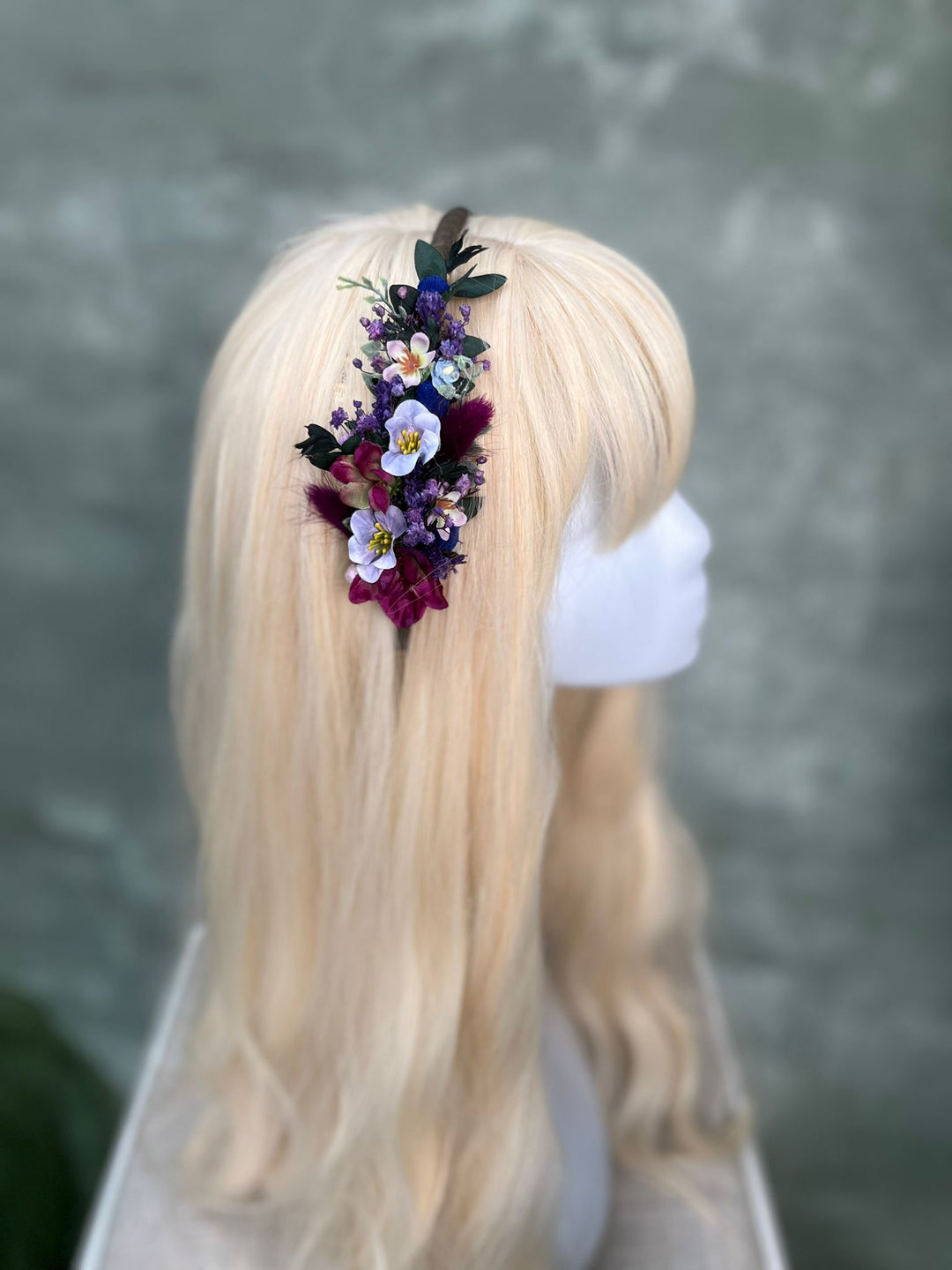 A blonde wig displayed on a white mannequin head, featuring a side-swept bang and a floral hair accessory with purple, blue , and dark blue flowers and green leaves.