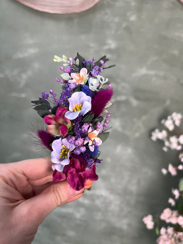 A hand holding a headband with purple, pink, and white blossoms, set against a soft gray textured background.