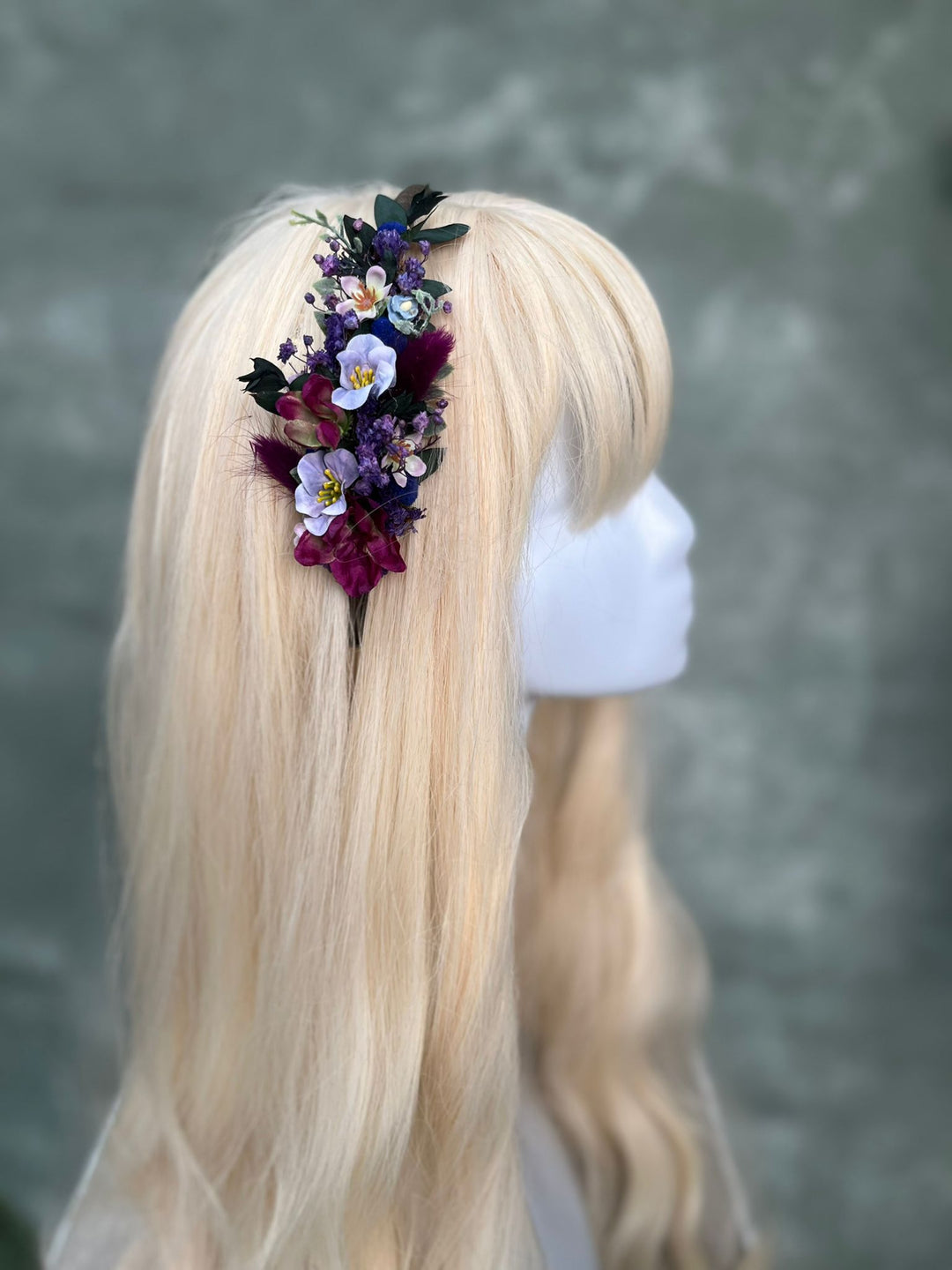A blonde wig displayed on a white mannequin head, featuring a side accessory of dried purple, white, and dark purple flowers with green leaves.