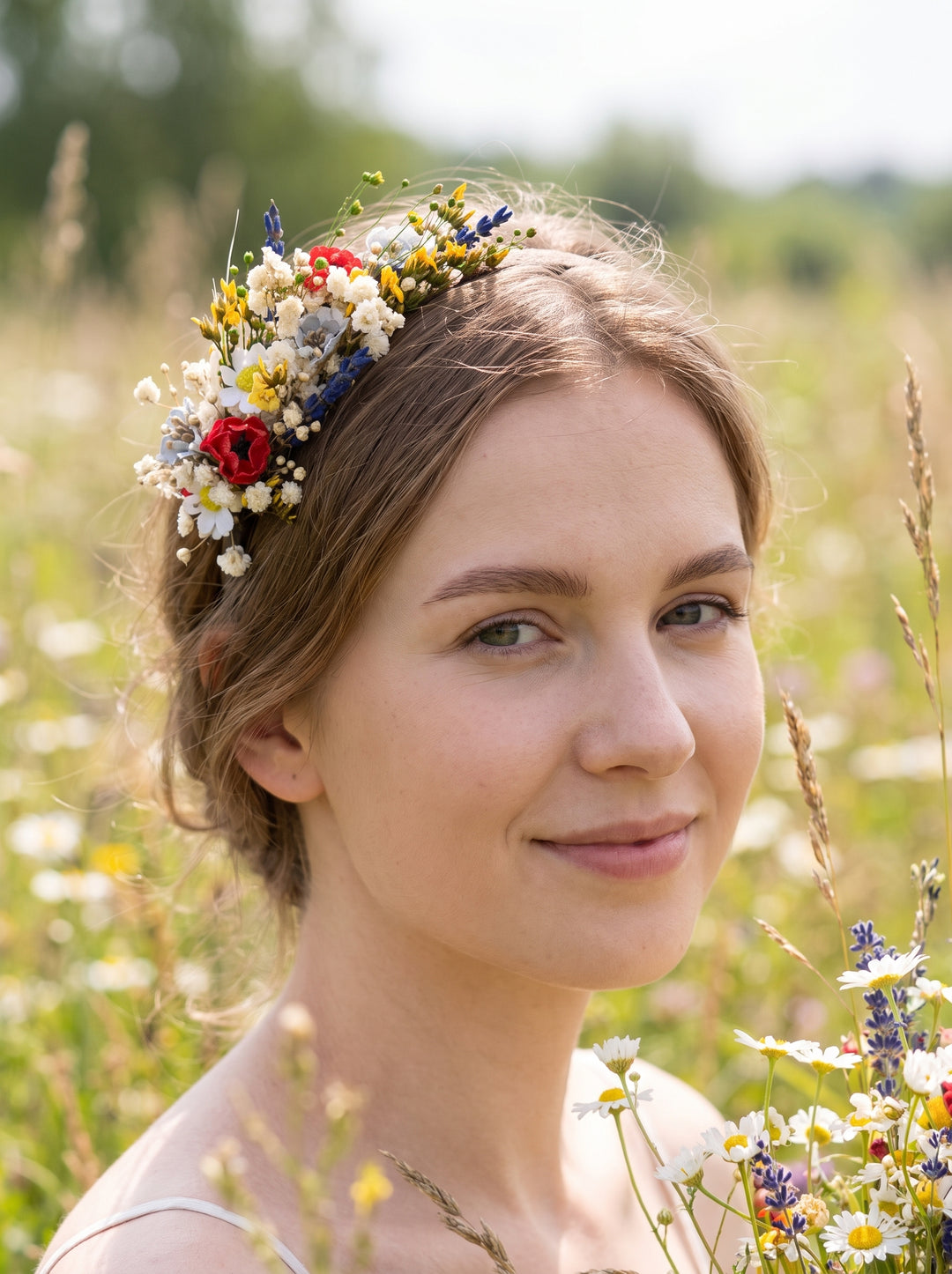 A woman outdoors in a sunlit field wearing a floral crown made of small white, yellow, blue, and red flowers. The face is obscured by a rectangular privacy block.