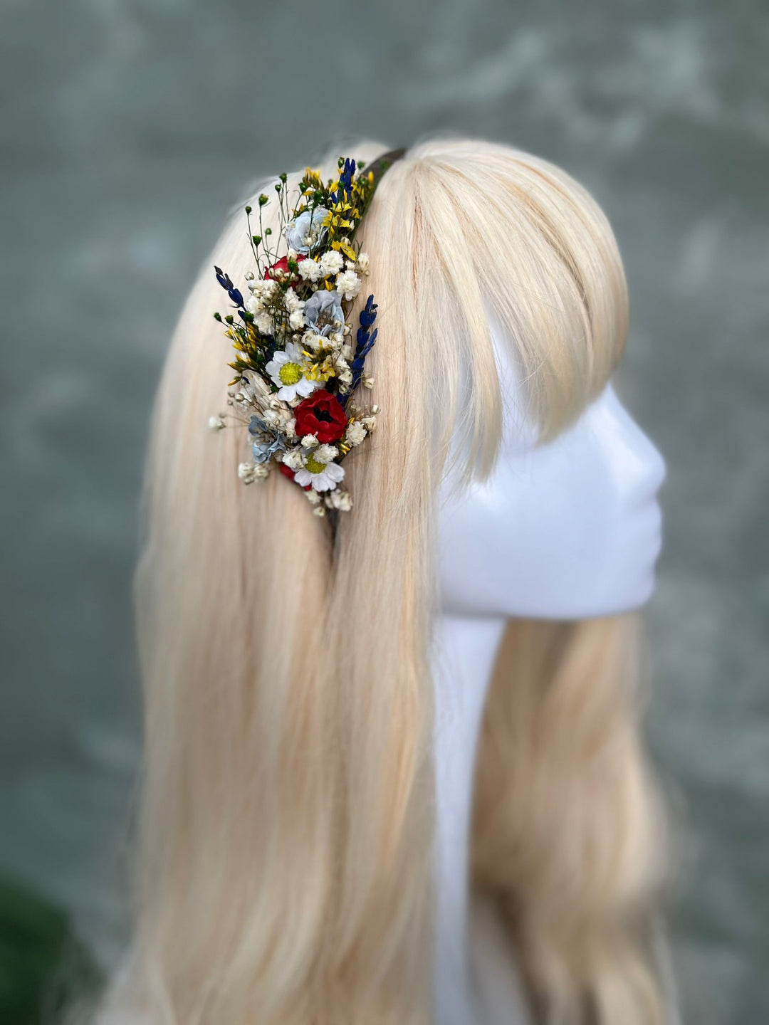 A blonde wig displayed on a white mannequin head, featuring a side accessory of dried flowers and small wildflowers.