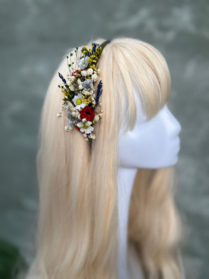 A blonde wig displayed on a white mannequin head, featuring a side accessory of dried flowers and small wildflowers.