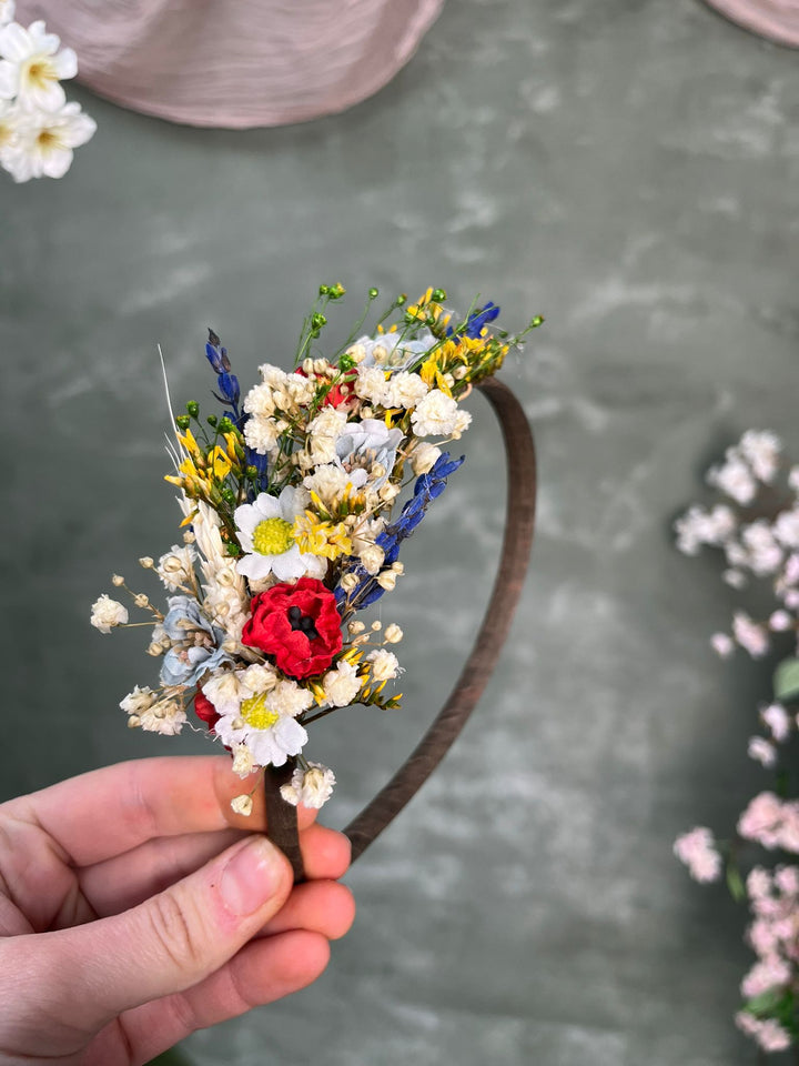 A hand holding a dark brown headband decorated with flowers in white, yellow, blue, and red tones, set against a textured gray-green background.
