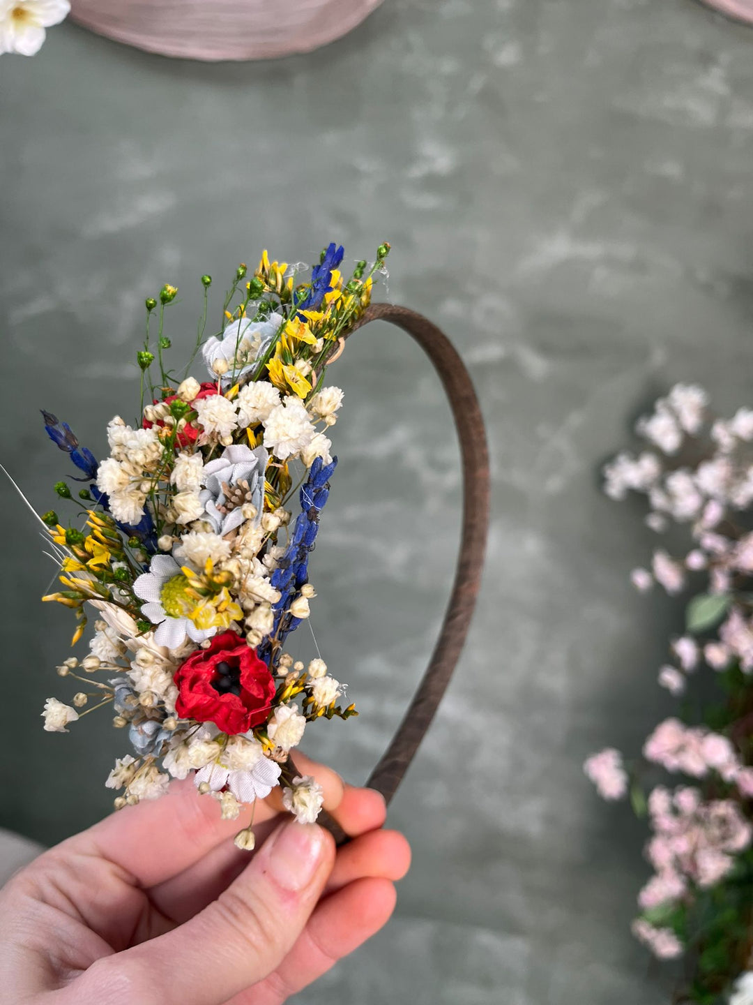 A hand holding a rustic brown headband decorated with a dense, colorful arrangement of wildflowers, including white, yellow, blue, and red blossoms, against a textured gray background.