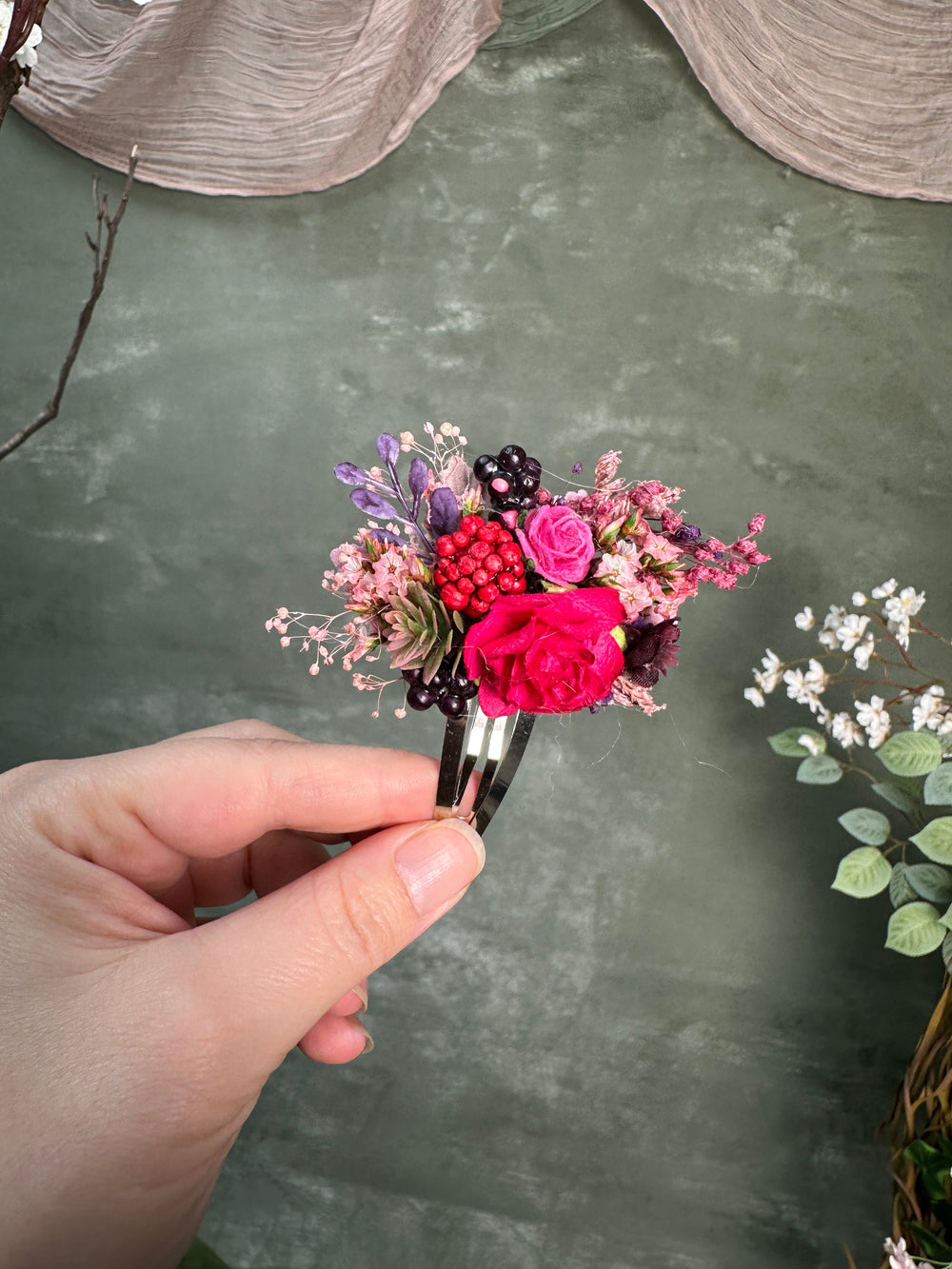 A hand holding a small flower hair clip with vibrant pink, red, purple, and black flowers nd berries, set against a textured dark green background.