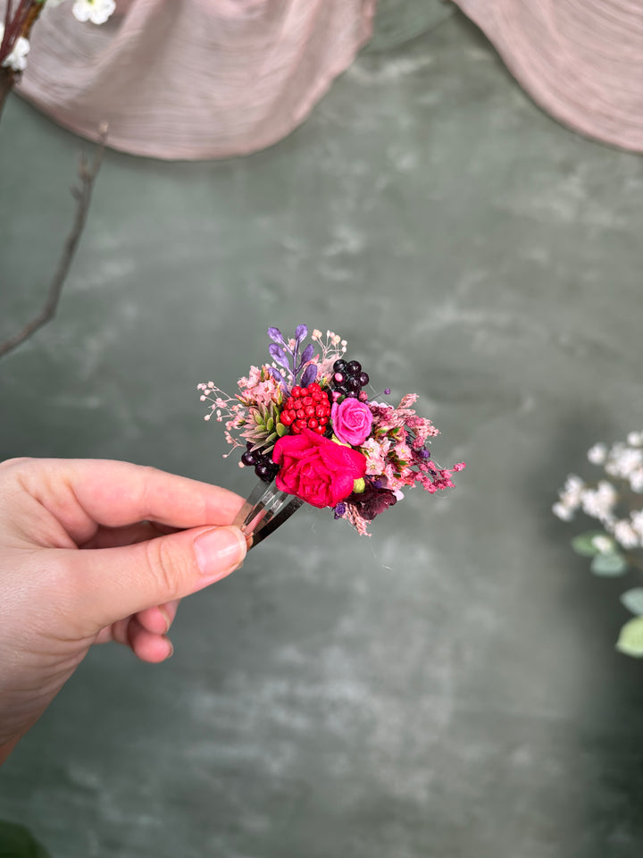 A hand holding a small dried flower hair clip with vibrant pink, red, purple, and dark berries against a textured dark green background.