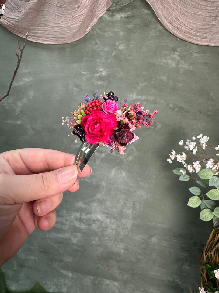 A hand holding a small floral hair clip with a vibrant pink rose and mixed dried and silk flowers, set against a textured gray-green background with decorative fabric and eucalyptus.