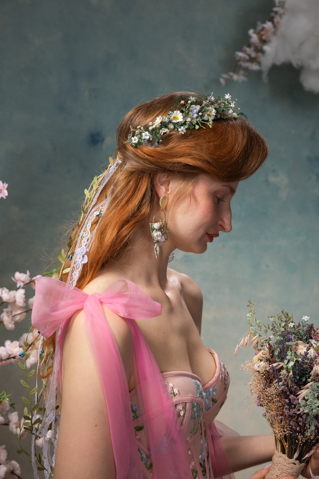 Woman with floral headpiece and pink fabric, holding flowers against a soft, blurred background