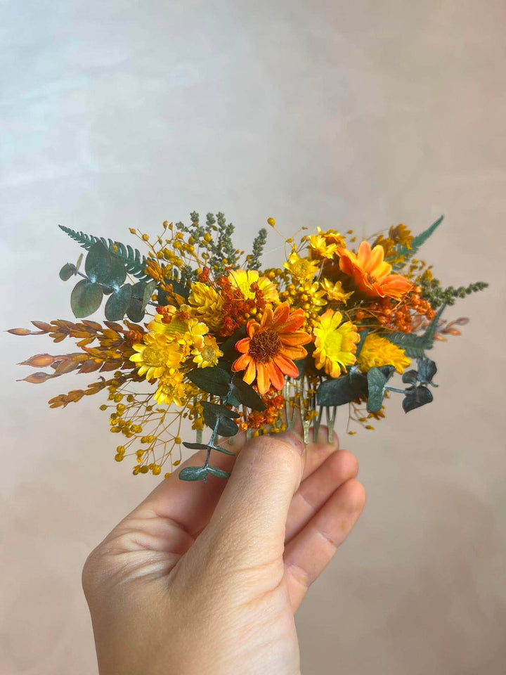 A hand holding a small flower hair comb with orange and yellow flowers and green foliage against a soft beige background.