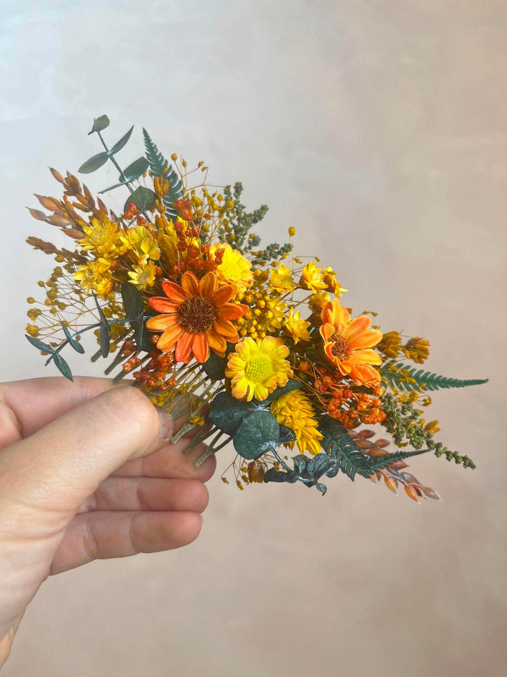 A flower hair comb with orange and yellow flowers and green foliage, held by a hand against a plain beige background.