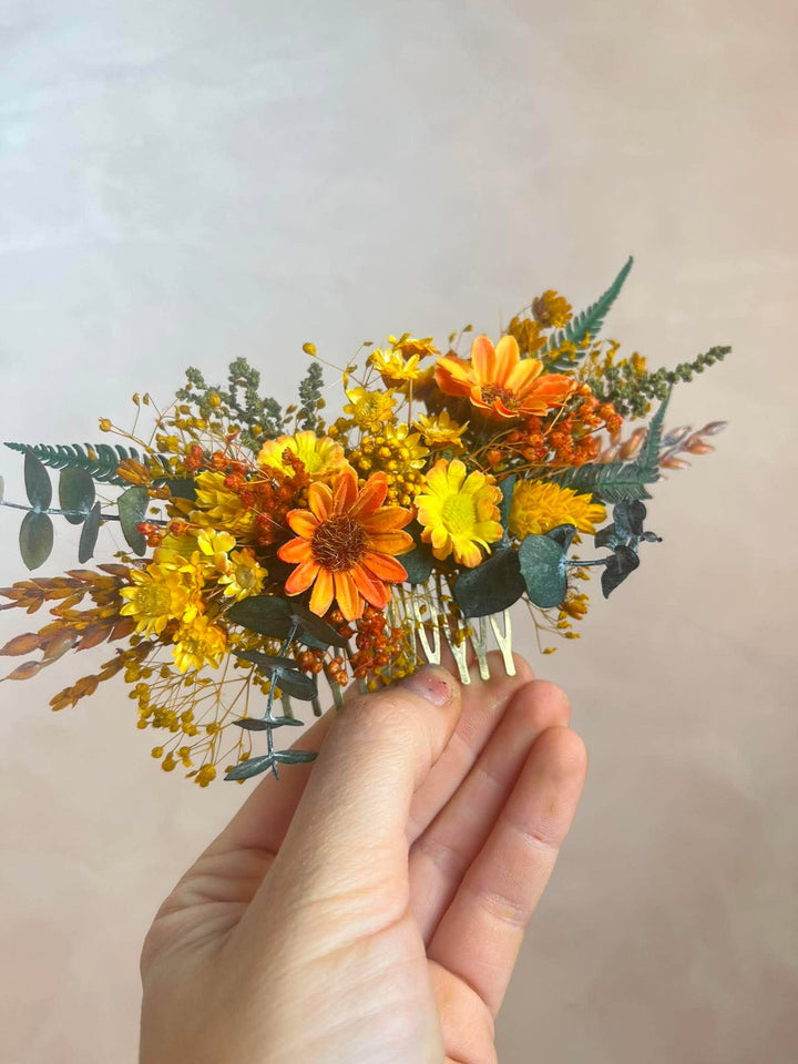 A hand holding a dried flower hair comb accessory featuring orange and yellow flowers with green foliage against a soft beige background.