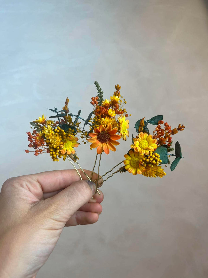 A hand holding a small flower hair accessory with yellow and orange flowers, green leaves, and gold-tone wire pins against a plain beige background.