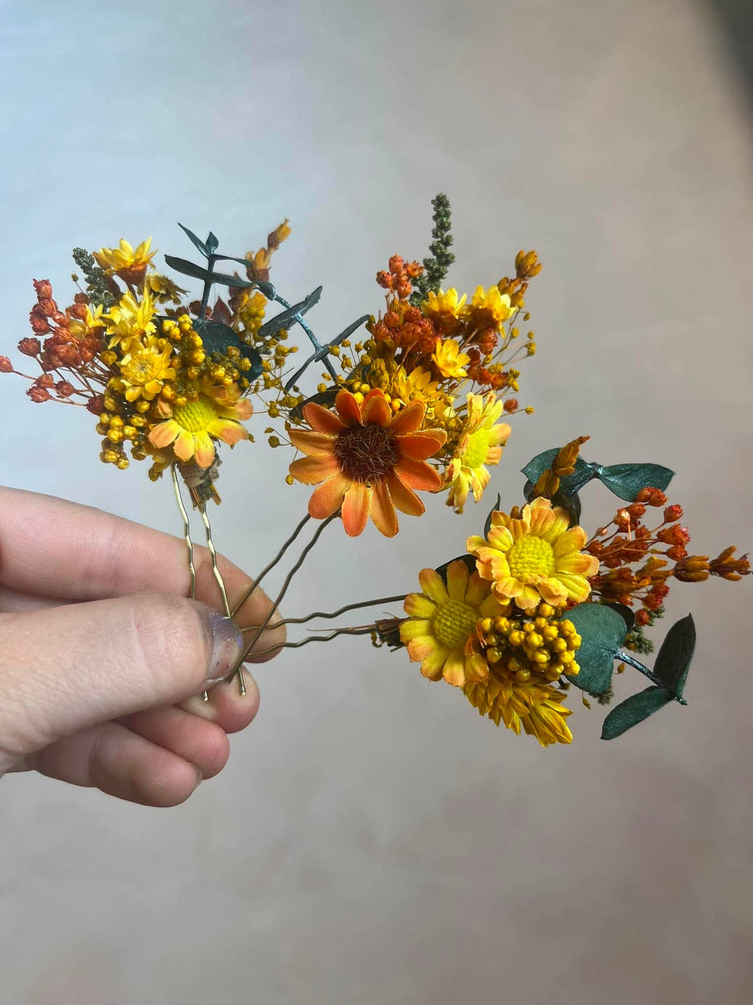 A hand holding a small hair floral arrangement featuring yellow and orange flowers with green and brown leaves against a plain light gray background.
